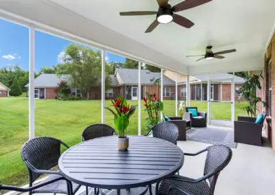 Screened-in porch sitting area with wicker furniture and red cushions, side table, lamp, wind chimes, and tray with wine bottle and glass