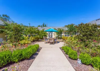 Courtyard with path leading to outdoor dining area surrounded by bushes, trees, and flower gardens