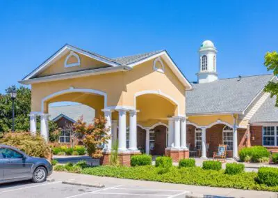 Large view of main entrance carport surrounded by grass, shrubs, and trees