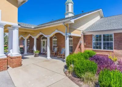 View of main entrance front porch area with rocking chairs next to grassy shrub/garden area