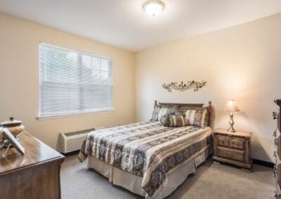 View of bedroom with bed, dresser, side table, lamp, large window, and various decor