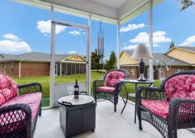 Screened-in porch sitting area with wicker furniture and red cushions, side table, lamp, wind chimes, and tray with wine bottle and glass